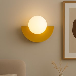 May include: A modern wall sconce featuring a bright, spherical light bulb set against a yellow, semi-circular backdrop. The fixture is mounted on a neutral-toned wall, creating a warm, inviting ambiance. The design is minimalist and contemporary.