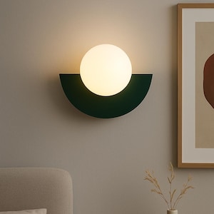 Op de afbeelding: Een moderne wandlamp met een halfronde donkergroene basis en een bolvormig, wit gloeiend licht. De armatuur is gemonteerd op een neutraal gekleurde muur, met een ingelijst kunstwerk en een vaas met gedroogde planten op de achtergrond.