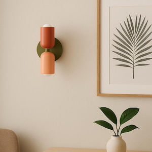 Könnte beinhalten: Eine moderne Wandleuchte mit zweifarbigem Design. Der obere Teil ist in einem gebrannten Orange gehalten, der untere Teil in Pfirsich. Die Leuchte ist auf einer runden, olivgrünen Basis montiert. Ein gerahmter botanischer Druck und eine kleine Vase mit Grün sind ebenfalls zu sehen.