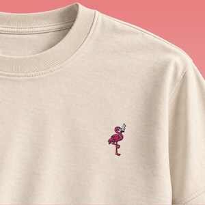 Könnte beinhalten: Ein beiges T-Shirt mit Rundhalsausschnitt und einer kleinen Stickerei auf der linken Brust. Das Motiv zeigt einen rosa Flamingo, der auf einem Bein steht und aus dessen Schnabel eine kleine weiße Rauchwolke aufsteigt, in detaillierter Stickoptik.