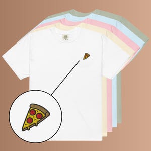 Könnte beinhalten: Ein Stapel kurzärmeliger T-Shirts in Weiß, Creme, Rosa, Hellblau und Salbeigrün. Jedes T-Shirt hat eine kleine gestickte Pizzascheibe auf der linken Brust. Ein kreisförmiger Einsatz zeigt eine vergrößerte Ansicht der Pizzastickerei.