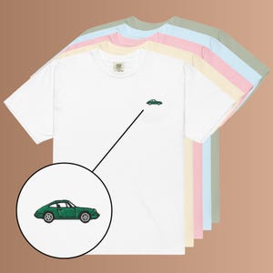 Könnte beinhalten: Ein Stapel kurzärmeliger T-Shirts mit Rundhalsausschnitt in Weiß, Gelb, Rosa, Hellblau und Salbeigrün. Jedes T-Shirt zeigt ein kleines, detailliertes grünes Sportauto, das auf der linken Brust gestickt ist, mit einer kreisförmigen Lupe.