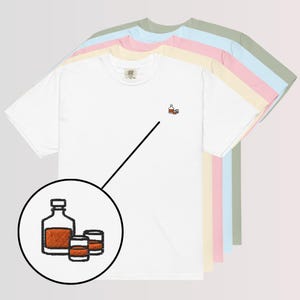 May include: White t-shirt featuring an embroidered design of a liquor bottle and two shot glasses in orange and brown. The shirt is shown with other pastel-colored t-shirts.