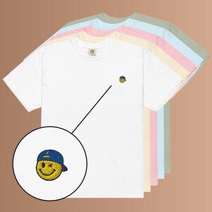 Könnte beinhalten: Ein Stapel kurzärmeliger T-Shirts mit Rundhalsausschnitt in Weiß, Creme, Rosa, Hellblau und Salbeigrün. Jedes T-Shirt hat eine kleine Stickerei auf der linken Brust, die ein gelbes, zwinkerndes Smiley-Gesicht mit blauer Baseballkappe zeigt.