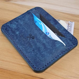 May include: A navy blue leather card holder with a distressed finish and visible stitching along the edges. The front features a small embossed logo of a hammer and wrench. A blue and white bank card and a portion of a bill are inserted.