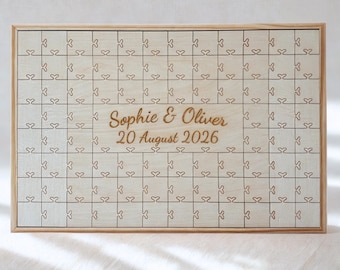 May include: A wooden puzzle guest book with a light wood frame. The puzzle pieces are etched with a heart design. The names "Sophie & Oliver" and the date "20 August 2026" are engraved in the center. A unique wedding keepsake.