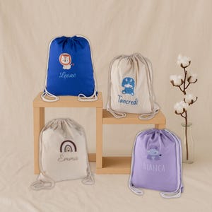 Könnte beinhalten: Vier Kordelzugbeutel in Blau, Beige und Lavendel. Jeder Beutel hat ein anderes gesticktes Design: einen Löwen, einen Dinosaurier, einen Regenbogen und ein Nilpferd. Die Beutel haben auch gestickte Namen: Leone, Tancredi, Emma und Bianca.