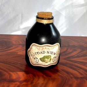 May include: A black glass bottle with a cork stopper and a label that reads "TOAD STEW." The label features a green illustration of a toad. The bottle is decorated with twine around the neck.