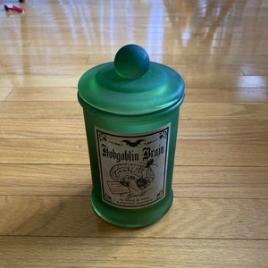 May include: A green glass jar with a frosted finish and a round lid. The jar has a label that reads "Hobgoblin Brain Spell to bind & heal a painful memory".