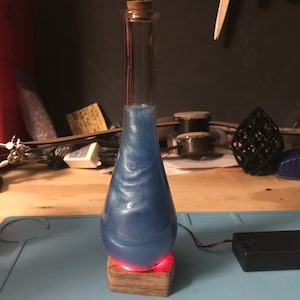 May include: A clear glass bottle with a cork stopper filled with a swirling blue liquid. The bottle is sitting on a wooden base with a red light underneath.
