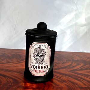 May include: Black glass jar with a black lid and a white label that says "Voodoo Potion". The label features a skull with a snake wrapped around it.