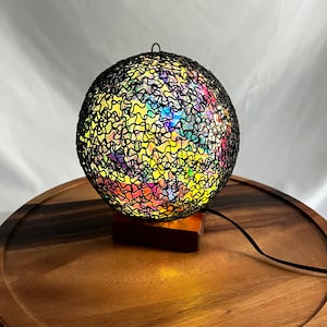 Puede incluir: Una lámpara de globo de alambre negro con un interior iridiscente de colores. La lámpara está sobre una base de madera y tiene un cable negro.