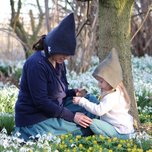 May include: Two people wearing pointed hats sit in a field of white flowers. The person in the blue hat is holding a leaf. The person in the green hat is looking down at the leaf.