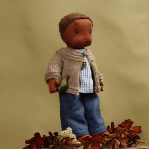 May include: A brown-skinned doll wearing a brown knitted cardigan, a blue and white plaid shirt, and blue jeans. The doll is holding a small green pinecone in its hand. The doll is standing on a bed of brown and green foliage.
