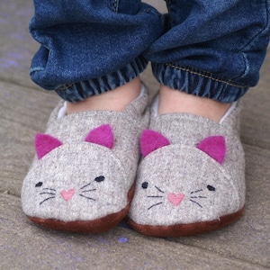 Puede incluir: Un par de pantuflas grises con forma de gato, con orejas de fieltro rosa y detalles faciales bordados. Las pantuflas las lleva puestas una persona que lleva pantalones vaqueros azules.