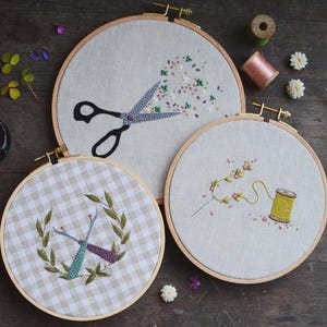 May include: Three embroidery hoops with floral and sewing-themed designs. One hoop features scissors with a floral burst, another has a wreath with sewing tools, and the third displays a floral vine and spool of thread. The hoops are set on a wooden surface.