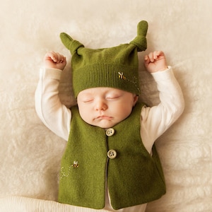 May include: A sleeping baby wearing a green knitted hat with bee embroidery and a green knitted waistcoat with bee embroidery.