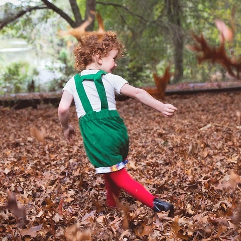 May include: A young child with curly red hair wearing a green overall with red tights and black shoes runs through a pile of autumn leaves.