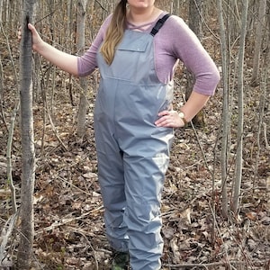 May include: A pair of grey waterproof overalls with adjustable straps. The overalls are being worn by a person standing in a woodland setting.