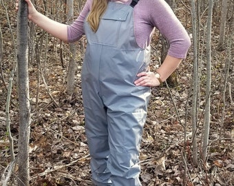 May include: A gray pair of waterproof overalls with adjustable straps. The overalls are worn by a person standing in a wooded area.