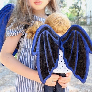 Könnte beinhalten: Ein Kind in einem blau-weiß gestreiften Kleid hält eine Puppe mit blauen und schwarzen Flügeln. Die Flügel sind aus Filz und haben einen glitzernden Überzug.