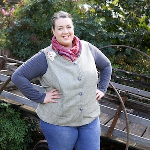 Puede incluir: Una mujer con un chaleco gris con un adorno floral, una bufanda roja y blanca y unos vaqueros azules. Está de pie delante de una rueda de carro de madera.