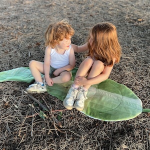 May include: Two children sit on a green and white leaf-shaped rug. The girl is wearing a white tank top and has long brown hair. The boy is wearing a white tank top and has short brown hair. They are both looking at each other.