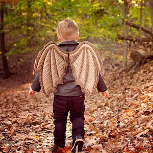 May include: A young child wearing a brown leather jacket and blue jeans walks through a woodland with large, brown, dragon wings attached to their back.