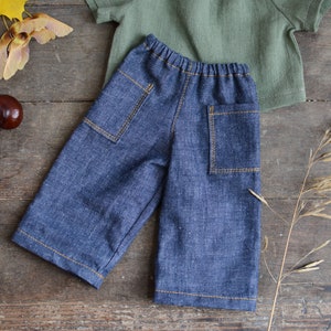 May include: Blue denim pants with two back pockets and a drawstring waist. The pants are laid flat on a wooden surface.