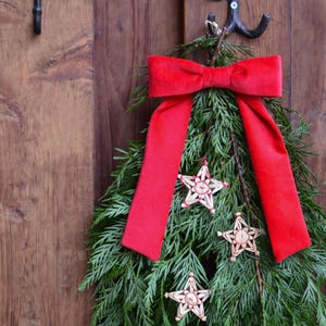 Könnte beinhalten: Ein festlicher Weihnachtskranz mit grünem Laub, einer großen roten Samtschleife und drei sternförmigen Ornamenten. Der Kranz hängt an einer Holztür und sorgt für eine warme und einladende Weihnachtsdekoration.