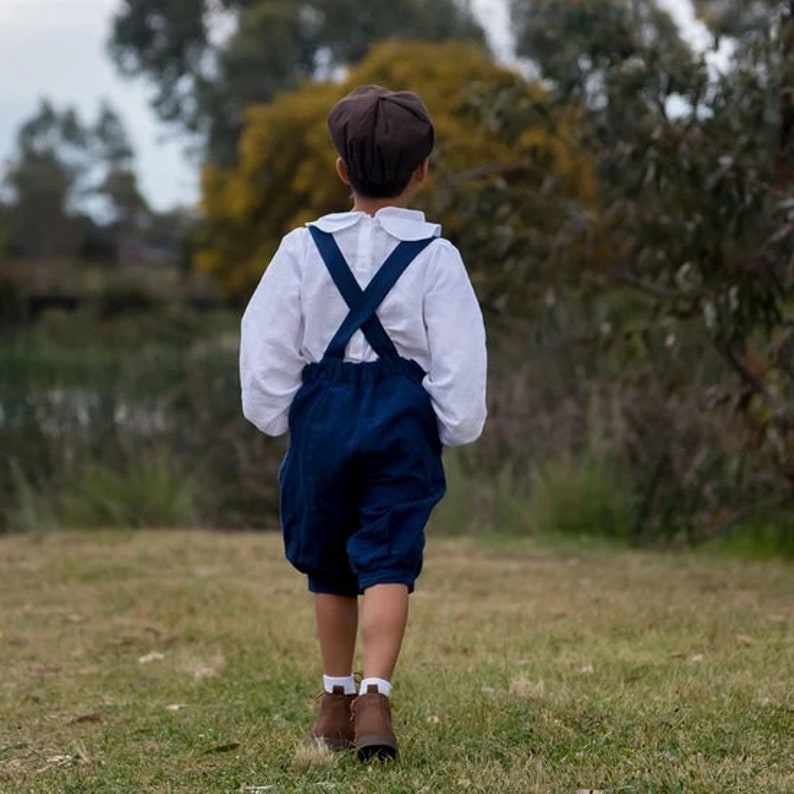 May include: A young person wearing a white collared shirt with a dark blue overall-style outfit and a brown cap. The person is walking on a grassy field.