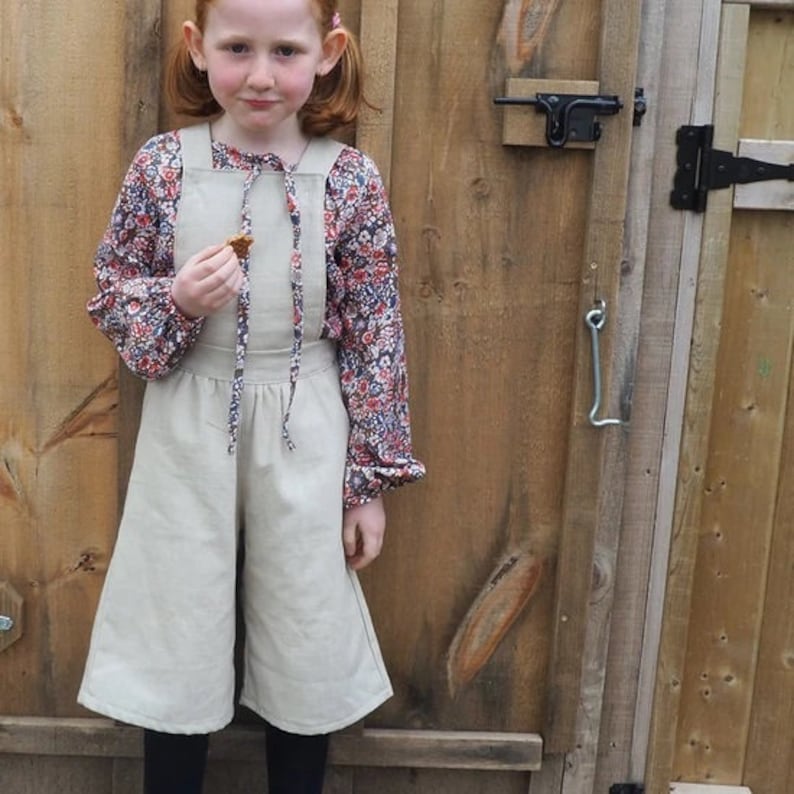 May include: A young person wearing a beige overall dress with a floral print blouse. The dress has a gathered waist and wide legs. The person is holding a small snack in their hand.
