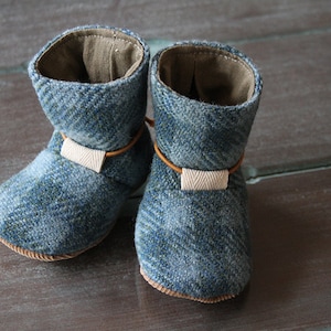 May include: A pair of baby booties made of blue tweed fabric. The booties have a light brown leather strap and a beige fabric accent. The interior is a light brown fabric. The soles are a light brown color.