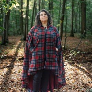 May include: A tartan cape featuring a pattern of red, dark blue, and green lines. The garment has a single button closure at the neck, an open front design, and a high-low hemline. It is worn over a dark long-sleeved shirt and dark trousers.