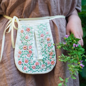 Puede incluir: Un delantal blanco con un diseño de bordado floral con rosas rosas y vides verdes. El delantal se ata a la cintura con una cinta color crema. El delantal se usa sobre un vestido marrón.