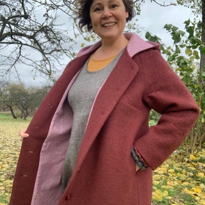 May include: A burgundy hooded coat with a pink lining. The coat has a button closure and pockets. The person wearing the coat is standing in a field of fallen leaves.