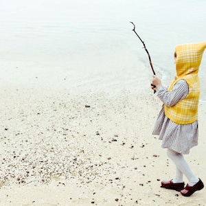 May include: A young person wearing a yellow and white plaid hooded vest and a plaid skirt, holding a stick on a sandy beach.