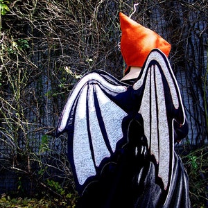Peut inclure: Un costume de chauve-souris noir et blanc avec un grand chapeau orange pointu. Les ailes sont en tissu avec un motif blanc et sont attach&eacute;es &agrave; une robe noire.