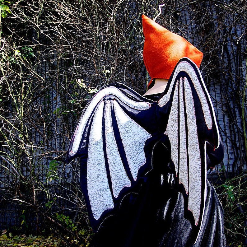 Peut inclure: Un costume de chauve-souris noir et blanc avec un grand chapeau orange pointu. Les ailes sont en tissu avec un motif blanc et sont attach&eacute;es &agrave; une robe noire.