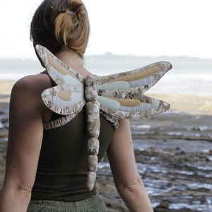 May include: A woman wearing a brown and green dragonfly costume with wings made of fabric. The wings are patterned with flowers and gold accents.