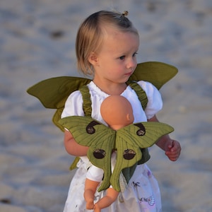 Può includere: Una bambina indossa un costume da falena verde e marrone e tiene una bambola. Il costume ha le ali e una cinghia che passa sopra le spalle della bambina. La bambina indossa un vestito bianco con un motivo floreale e un design di unicorno.