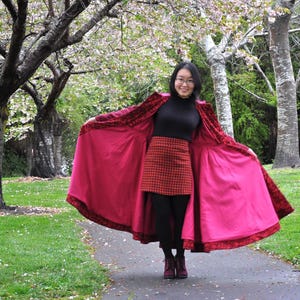 May include: A person wearing a red velvet coat with a bright pink lining, a black turtleneck, and a red plaid skirt. The coat has ornate trim and is open, revealing the lining. The person is standing on a path in a park with trees and flowers.