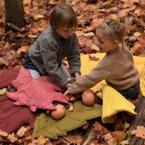 May include: Two children play with dolls on a colorful leaf-shaped play mat. The mat is made of felt and has a red, pink, and green color scheme. The children are wearing casual clothing and are sitting on the floor.