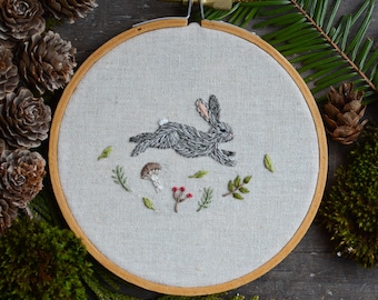 May include: A gray embroidered rabbit hops through a forest scene on a white linen fabric stretched in a wooden embroidery hoop.