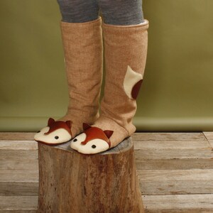 May include: A pair of brown knee-high boots with fox faces on the toes. The boots are made of a soft, fuzzy material and have a brown and white color scheme.