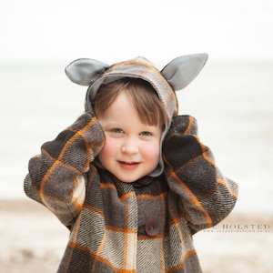 May include: A young child wearing a plaid coat with a gray hooded hat with ears. The child is smiling and looking at the camera.
