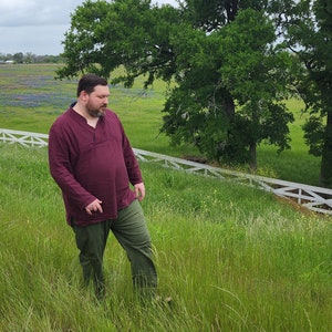 May include: A man wearing a maroon long-sleeved shirt and green trousers walks through a field of wildflowers. He is walking towards a white wooden fence.