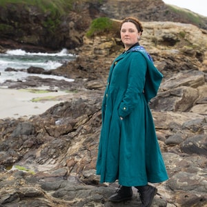 May include: A teal-colored hooded coat with gold buttons and pockets. The long coat is worn outdoors near a rocky beach. The coat has a full skirt and a high collar. The coat is made of a textured fabric.