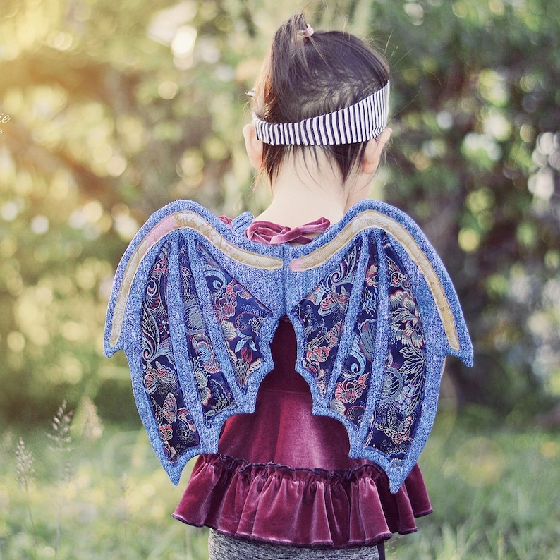 Peut inclure: Une enfant portant une robe en velours violet avec un ourlet &agrave; volants et une paire d'ailes de dragon bleues et dor&eacute;es. Les ailes sont en tissu et ont un motif floral.