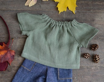 Puede incluir: Una blusa de lino verde con cuello fruncido y mangas cortas. La blusa está sobre una superficie de madera con un par de pantalones vaqueros azules y decoraciones de otoño.